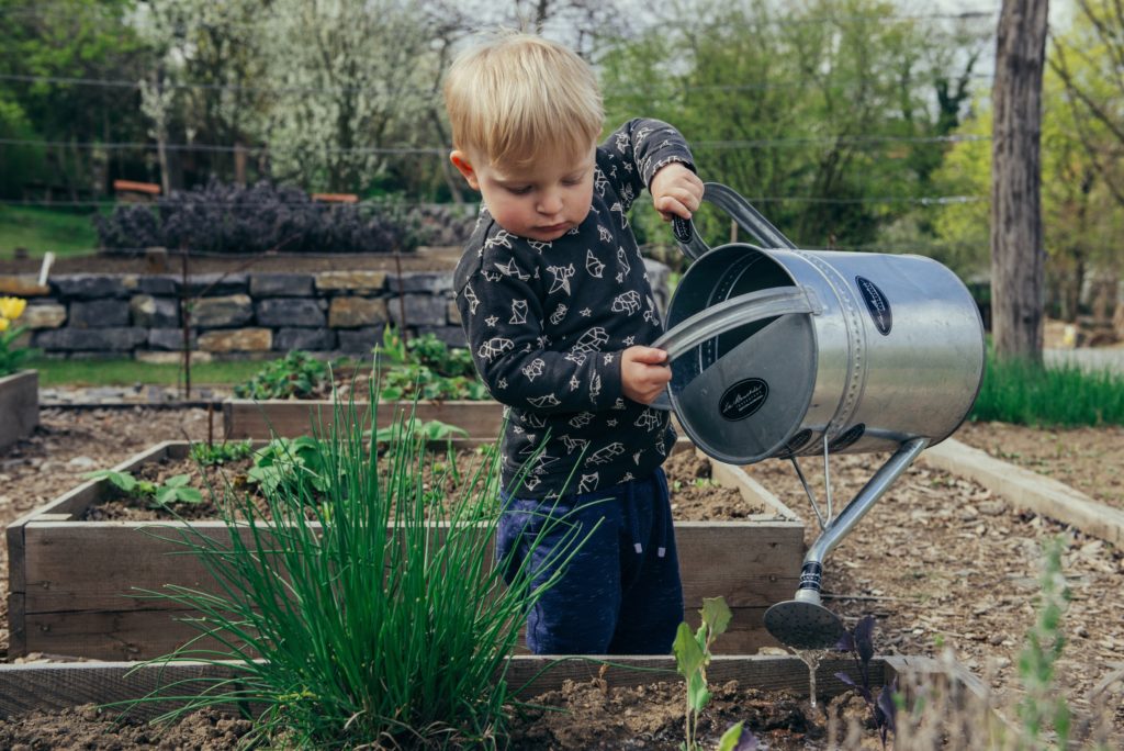 jeune garçon en manche longue noir tenant un arrosoir métallique pour arroser les bacs d'herbes aromatiques